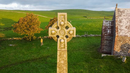 Aerial view of large outdoor Celtic cross, standing in a green graveyard next tocountry chapel...