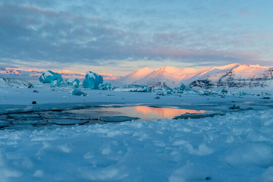Icebergs and huge chunks for green-blueish ice fill Jokulsarlon Glacier lagoon, as the golden hour sunlight of the early morning casts a beautiful warm glow over the glacier-filled national park in - Powered by Adobe