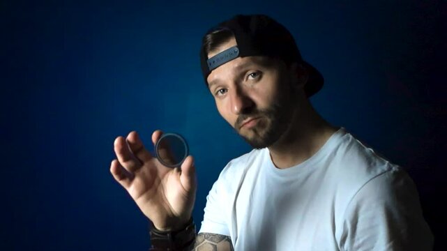 Man with white shirt black reversed cap hat and tattoos holds up a CPL polarizer filter and looks through it with confidence and happiness in slow motion while being in front of a blue studio wall.
