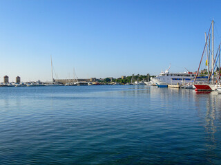 A tranquil view of the harbor in Rhodes, Greece, featuring historic windmills, sailing yachts, and...
