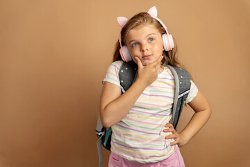 Portrait of a school-age kid in headphones with a school backpack thoughtfully looking aside on a...