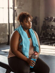 A woman sits in the gym after a workout with a bottle of water and a towel