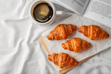 French croissants, mug of coffee and opened book beside on white bedding linen. Top view, flat lay, copy space, horizontal orientation. Meal serve. Breakfast in bed,  tasty morning concept