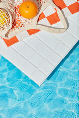Summer beach towel with orange checks, fresh fruit, and sunscreen tube styled on white tiles beside...
