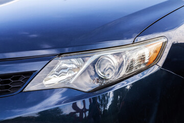 The headlight of a car is lit up. The car is blue. The sky is cloudy