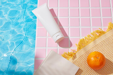 A soft beach towel and tangerine lie atop pink mosaic tiles beside a shimmering pool. A blank tube...