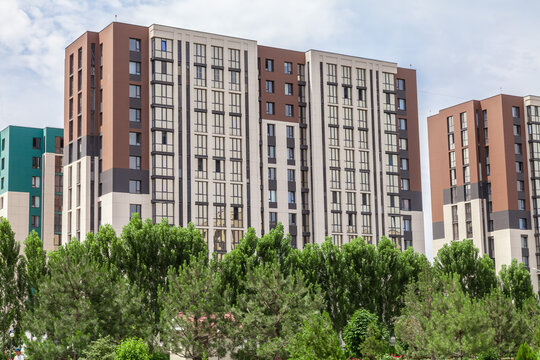 A city skyline with tall buildings and a large arch. The buildings are mostly brown and white, and the arch is a curved shape. The sky is cloudy, and the trees are green and lush