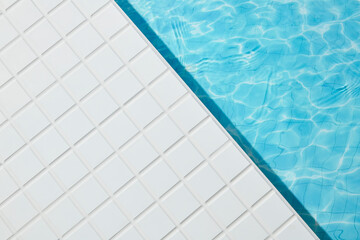 Empty poolside shot with textured white mosaic tiles and calm water reflection. No objects present,...