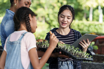 Asian teenage student holding vegetable tray while smiling at african female teacher and asian preteen girl during outdoor organic farming lesson at school garden with teamwork and technology tools