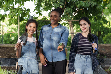 Multiracial group of female students and african teacher smiling with gardening tools in hand, standing together in outdoor school agriculture class with soil beds and trees in natural environment