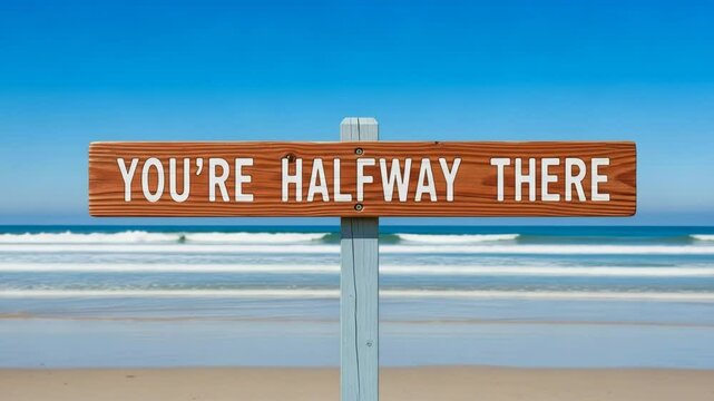 A wooden sign on a beach reads youre halfway there, offering encouragement and symbolizing progress on a journey
