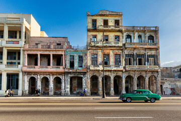 Havana, Cuba - 07 July 2025: View of aged, pastel-hued buildings stand proudly against the clear sky, while a vintage green car cruises along the sun-drenched street.