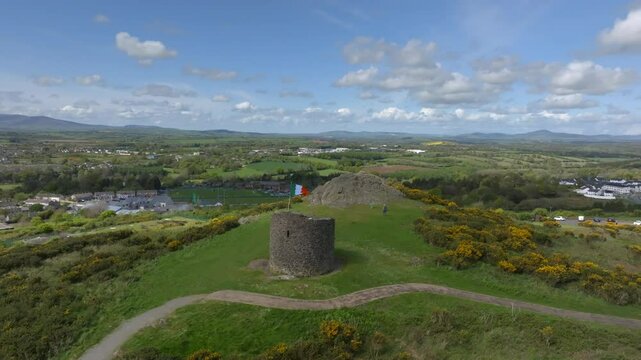 Vinegar Hill, Enniscorthy, County Wexford, Ireland, April 2025. Drone orbits clockwise slowly pulling back following winding dirt trail around monument tower with Irish flag rippling in wind.