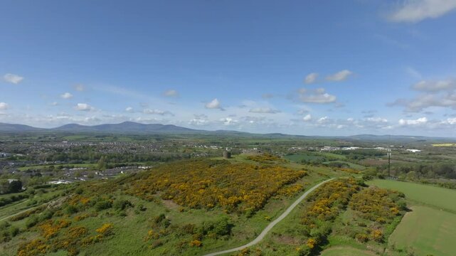 Vinegar Hill, Enniscorthy, County Wexford, Ireland, April 2025. Drone wide overview orbits around dense shrub low brush growing up along to stone tower monument under speckled cloudy sky.