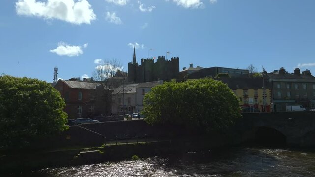 Enniscorthy, County Wexford, Ireland, April 2025. Drone low tracking upstream along River Slaney pulls back to ascend over road with town centered and cars crossing bridge leaving village.
