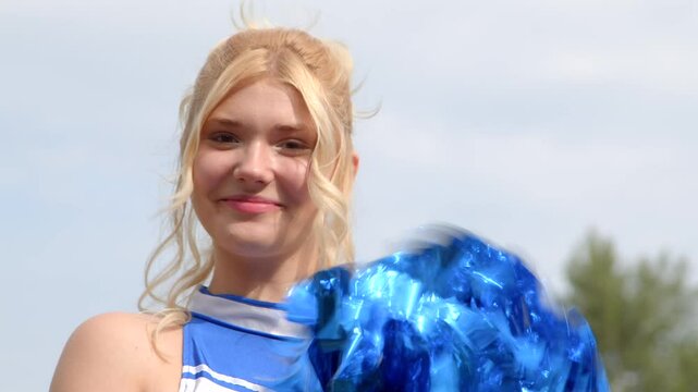 Close-up of positive attractive high school cheerleader in uniform with bright pompoms making clap cheer motions, looking with cheerful smile, expressing excitement and joy outdoors.