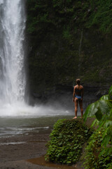 Woman standing on mossy rock gazing at jungle waterfall in Bali