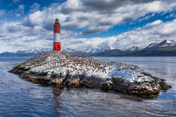 View of the stark red and white Les Eclaireurs Lighthouse stands sentinel on a rocky islet teeming with birds, set against snow-capped mountains, Ushuaia, Tierra del Fuego Province, Argentina.