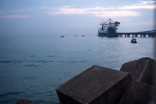 Cilegon, Indonesia-September 14, 2023: Container ship docked at a port with cranes and a pier, small boats in the foreground, hazy industrial sea view.