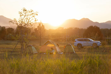 A heartwarming camping scene during golden hour with tents, a car, and silhouettes of people enjoying nature under a warm sunset in the open field.