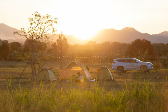A heartwarming camping scene during golden hour with tents, a car, and silhouettes of people enjoying nature under a warm sunset in the open field.