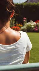 Woman sitting outdoors with her back to the camera, enjoying the sunny day in a beautiful green garden with flowers.