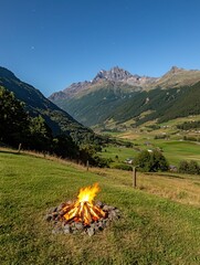 A campfire burns on a grassy hill overlooking a valley with scattered houses, surrounded by mountains under a clear starry night sky.