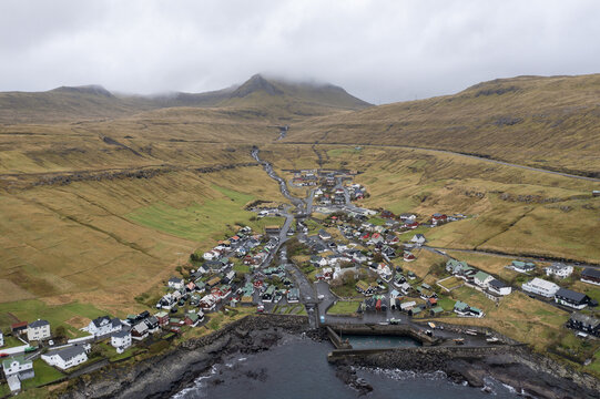Aerial view of a quaint village nestled between dramatic green hillsides and dark rocky coastlines under a moody, overcast sky, Tj&Atilde;&cedil;rnuv&Atilde;&shy;k, Streymoy, Faroe Islands.