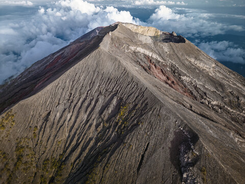 Aerial view of the imposing Agung volcano, its rugged slopes etched with deep ravines, standing tall amidst a sea of fluffy clouds, Karangasem, Bali, Indonesia.
