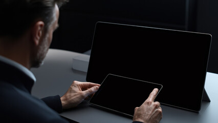 Businessman sitting at minimalist desk reviewing digital tablet and large blank monitor, modern technology workspace, professional, focused, contemporary office environment