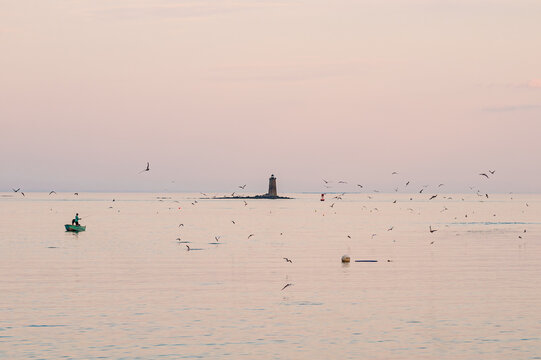 Lone boat on calm sea with lighthouse and birds at sunset.