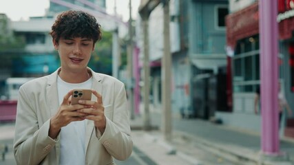 Portrait of smiling male office employee typing messages on smartphone walking along city district. Businessman communicates with colleagues in work chat on street. Mobile technology - Powered by Adobe