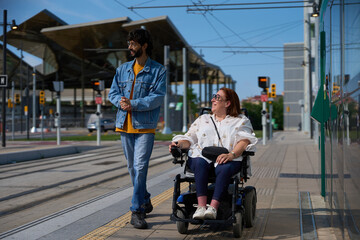 Man walking with woman using electric wheelchair at tram stop - wheelchair and disability concept