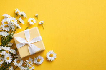 Gift box with white ribbon and bunch of Chamomile flowers on bright yellow background, top view, copy space. Mothers Day, Birthday, Anniversary celebration concept template