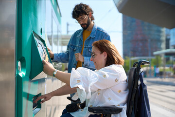 Woman in wheelchair using ticket machine with friend at tram or train station - wheelchair and...