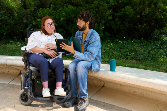 University student helping classmate in wheelchair studying in park - wheelchair and disability concept
