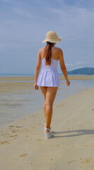 Stunning Asian woman in a short white dress and straw hat strolls confidently along a sunny beach