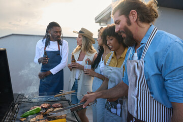 Friends grilling meat and vegetables, enjoying drinks on rooftop at sunset