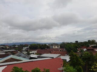 A cloudy sky over a residential area with red rooftops, green trees, and distant mountains visible...
