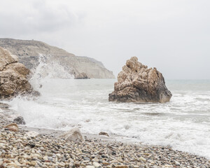 View of waves crashing against rocky outcrops and pebble beach on a grey day, the sea mist blending into the distant cliffs, Paphos, Paphos, Cyprus.