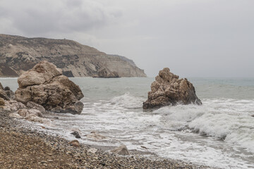 View of waves crashing against imposing rocks on a pebble-strewn beach under a grey sky, as a rugged coastline rises in the background, Paphos, Paphos, Cyprus.