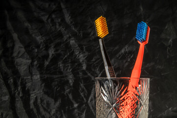Two toothbrushes, one orange and one black, stand upright in a crystal glass on a dark background. The contrast of colors and textures creates a striking visual