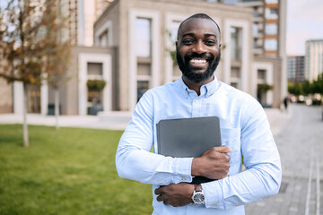 Smiling businessman stands confidently with laptop in hand outside modern office building in urban...