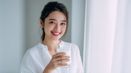 Woman holding a glass of milk while smiling in a bright, sunlit room with sheer curtains during the morning