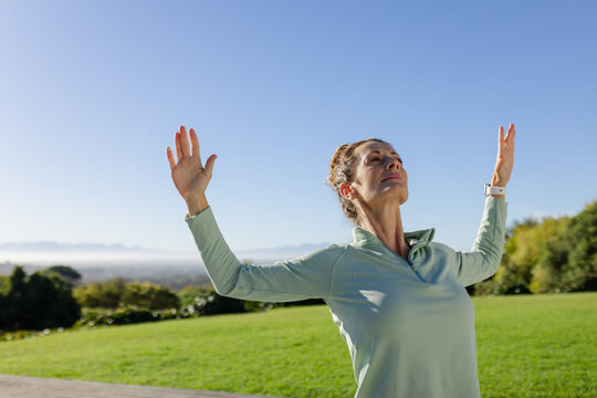 Female stretching in park raising arms upward wearing green quarter-zip top, white fitness tracker