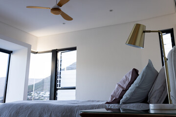 Bed with gray bedding and pillows sitting beside brass lamp and fan near black-framed windows