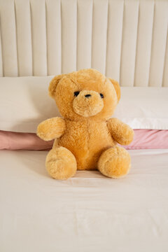 Plush teddy bear is resting on white bed sheets beside pale pink pillow, cream tufted headboard