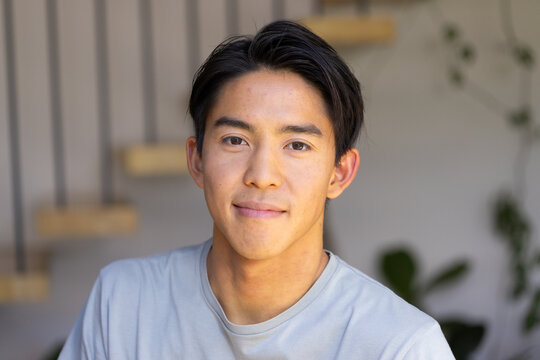 Asian man standing indoors by floating stairs with railings facing camera wearing grey tshirt