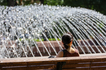 Woman sitting on a bench against the fountain splashes in summer park. Hot weather in city