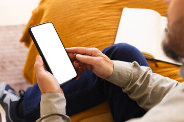 Asian man sitting on couch at home holding smartphone with blank screen next to open notebook